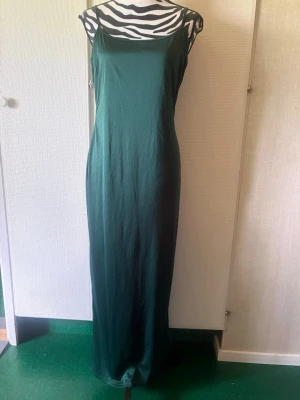 Grön långklänning med smala axelband - Elegant grön långklänning med smala axelband och rak siluett. Klänningen har en enkel och stilren design, perfekt för dig som gillar minimalistiskt mode. Tillverkad i ett glansigt material som ger en lyxig känsla.