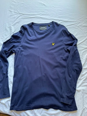 Marinblå långärmad tröja Lyle & Scott - Snygg marinblå långärmad tröja från Lyle & Scott med rund halsringning. Tröjan har det klassiska gula fågel-logotypen broderad på bröstet. Tillverkad i mjuk bomull, perfekt för en stilren och avslappnad look. Kan gå ned i pris vid snabb affär!