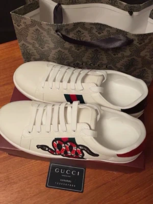 Gucci Ace med orm - Snygga vita sneakers från Gucci med klassiska röd-blå ränder på sidan och en broderad orm i rött, svart och vitt. Skorna har lågt skaft, vita snören och detaljer i läder. Perfekta för dig som vill sticka ut med lyxig streetstyle. Storlek 41 men passar ändå till 40. Priset är ej hugget i sten!