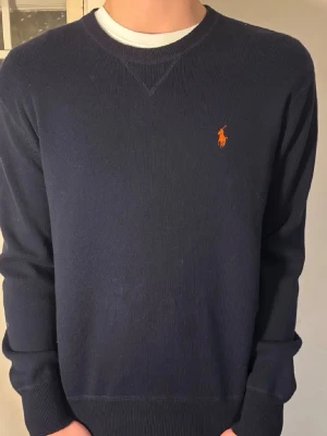 Mörkblå tröja från Polo Ralph Lauren - Klassisk mörkblå tröja från Polo Ralph Lauren med rund halsringning och den ikoniska orange logotypen broderad på bröstet. Tröjan har långa ärmar och ribbade muddar vid ärmslut och nederkant. Perfekt för en stilren och enkel look.