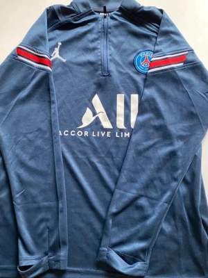 PSG träningsset från Jordan x Nike - Mörkblått PSG träningsset med långärmad tröja och byxor. Tröjan har dragkedja vid halsen, vita och röda detaljer på ärmarna samt PSG och Jordan-logga. Byxorna har elastisk midja, röda och vita ränder samt PSG och Jordan-logga. Perfekt för fotbollsträning.
