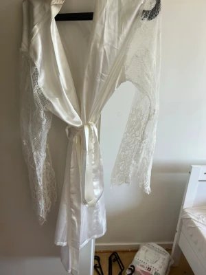 Vit satin morgonrock med spetsärmar - Snygg vit morgonrock i satin med långa ärmar i transparent spets. Knytband i midjan och glansig finish ger en lyxig känsla. Perfekt för att chilla hemma eller som elegant loungewear. Materialet är mjukt och följsamt, och spetsdetaljerna gör den extra speciell.