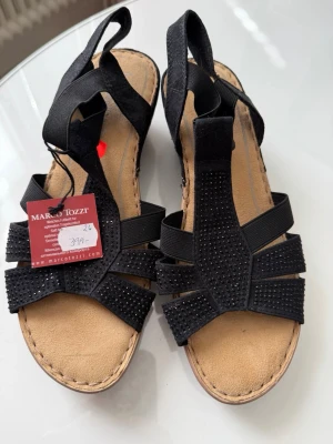 Svarta sandaler från Marco Tozzi - Stilrena svarta sandaler från Marco Tozzi med elastiska band och dekorativa svarta små stenar på remmarna. Mjukt beige innersula och öppen tå. Perfekta för en trendig och bekväm look under varma dagar.