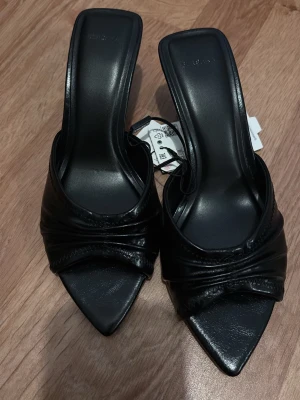 Svarta pumps med spetsig tåg  - Stilrena svarta pumps med öppen häl och fyrkantig tå. Skorna har en elegant klack och är tillverkade i ett glansigt material med snygga veckdetaljer framtill. Perfekta för dig som gillar trendiga och minimalistisk skor.