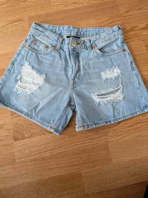 Ljusa denimshorts med slitningar - Säljer ett par ljusblå denimshorts med snygga slitningar framtill. Klassisk femficksmodell med knapp och dragkedja. Perfekta för sommaren och ger en avslappnad vibe.
