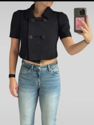 Svart croppad kavajjacka med knappar - Snygg svart croppad kavajjacka med korta ärmar och dubbelknäppning framtill. Jackan har klassisk krage och dekorativa axelpartier. Perfekt för en trendig och stilren look.