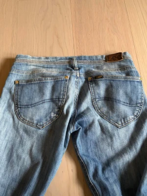 Blå jeans från Lee med raka ben - Säljer ett par klassiska blå jeans från Lee med raka ben och fem fickor. Jeansen har en ljus tvätt och tydliga sömmar på bakfickorna. Tillverkade i bomullsdenim och har Lee-logga på bakfickan.