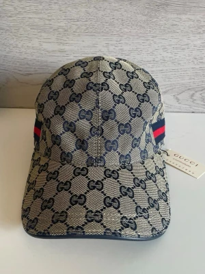 Gucci keps med GG-mönster och rand - Snygg keps från Gucci i beige och marinblått med klassiskt GG-monogram över hela kepsen. På sidan finns en ikonisk röd och marinblå rand. Tillverkad i canvasmaterial och har en böjd skärm. Perfekt för dig som vill ha en exklusiv och trendig accessoar.