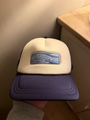 Blå och beige keps från Les Deux - Snygg keps från Les Deux i skum med blå skärm och beige frontpanel. Framsidan har ett blått broderat märke med texten 'Texas Hollywood'. Baksidan är i mesh för extra ventilation. Perfekt accessoar för en avslappnad stil.