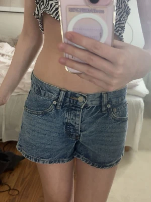 Blå denimshorts med broderade fickor - Säljer ett par klassiska blå denimshorts med snygga broderade detaljer på bakfickorna. Shortsen har hög midja, med fickor och bälteshällor. Perfekta till sommaren och lätta att matcha med olika toppar.