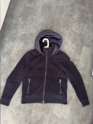 Moncler cardigan  - Snygg mörkblå stickad jacka från Moncler med quiltad huva och dragkedja framtill. Jackan har två fickor med dragkedja och ribbade muddar. Perfekt för dig som vill ha en stilren och bekväm jacka med exklusiv känsla.