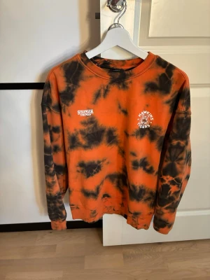 Stranger Things tie-dye tröja orange - Cool orange och svart tie-dye sweatshirt från Stranger Things med tryck 'Hawkins Tigers' och basketmotiv. Rund halsringning och långa ärmar. Perfekt för dig som gillar serien och vill sticka ut med en unik stil.