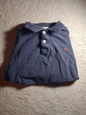 Mörkblå pikétröja med röd logga - Klassisk mörkblå pikétröja med krage och tre vita knappar framtill. Liten röd broderad logga på bröstet. Perfekt till jeans eller shorts för en avslappnad stil.