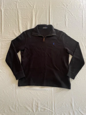Svart halvzip-tröja från Polo Ralph Lauren - Svart långärmad tröja med halv dragkedja från Polo Ralph Lauren. Tröjan har en klassisk krage och blå broderad logga på bröstet. Tillverkad i mjukt bomullsmaterial och har ribbade muddar vid ärmslut och nederkant.