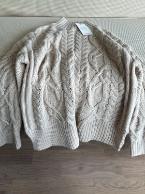 Beige kabelstickad tröja - Snygg beige kabelstickad tröja med grovt mönster och ribbade muddar. Tröjan har rund halsringning och är perfekt för lager-på-lager-stil. Materialet känns mjukt och värmande, perfekt för kyligare dagar.