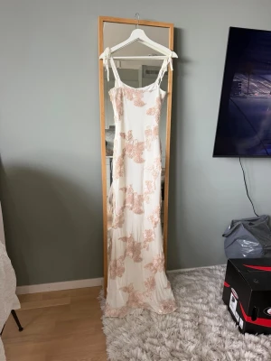 Lång vit klänning med blommiga detaljer - Elegant långklänning i vit med ljusbeige blommiga broderier. Klänningen har smala axelband med knyt och en figurnära passform. Perfekt för sommarens speciella tillfällen.