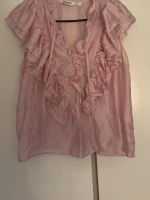 Rosa volangblus  - Söt ljusrosa blus i glansigt material. Blusen har volanger längs urringningen och axlarna samt knytband framtill. Ärmlös modell som ger en luftig och feminin känsla. Perfekt för att lyfta vilken outfit som helst.