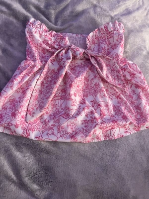 Rosa blommig bandeau topp med knyt - Supersöt bandeau topp i vitt med rosa blommigt mönster och stor knytning framtill. Toppen har volangdetaljer upptill och smockad rygg för extra komfort. Perfekt för en somrig och trendig look.
