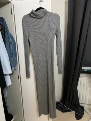 Grå ribbad poloklänning från MNG - Långärmad, figurnära klänning i grått från MNG. Klänningen är ribbstickad med polokrage och har en lång, slimmad siluett. Perfekt för dig som gillar en stilren och enkel look med lite extra edge.