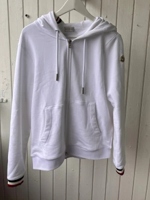 Vit hoodie med dragkedja från Moncler - Snygg vit hoodie från Moncler med dragkedja framtill, huva med dragsko och två stora fickor. Ärmsluten har coola ränder i svart och rött. Moncler-logga på ärmen. Mjuk bomullskänsla och perfekt för en clean streetstyle-look. Pris kan diskuteras vid snabb affär
