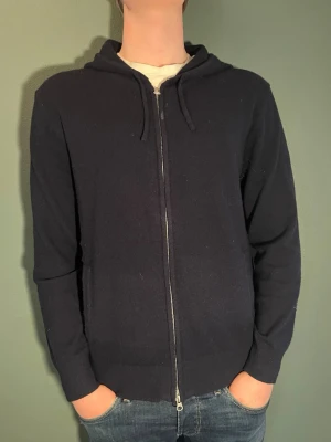 Mörkblå hoodie med dragkedja - En stilren mörkblå hoodie med dragkedja framtill och huva med dragsko. Tillverkad i mjukt material och har ribbade muddar vid ärmslut och nederkant. Perfekt för en avslappnad look och enkel att matcha med jeans eller joggers. Nypris ca 1000 kr