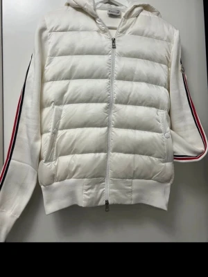 Vit Cardigan med huva från Moncler - Snygg Vit Cardigan från Moncler med huva och dragkedja framtill. Jackan har quiltad front, stickade ärmar med röda och mörkblåa ränder samt Moncler-logga på ärmen. Perfekt för kyliga dagar och ger en sportig vibe. Passar även S