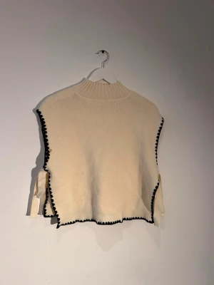 Beige stickad väst med svarta detaljer - Snygg beige stickad väst med svarta virkade kanter och sidosnörning. Hög ribbad krage och lös passform ger en trendig look. Perfekt att styla över en skjorta eller t-shirt för en cool lager-på-lager-effekt.