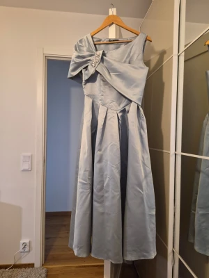 Ljusblå långklänning med rosett - Elegant ljusblå långklänning i satinliknande material med en axel och stor dekorativ rosett framtill, prydd med pärlor. Klänningen har veckad kjol och asymmetrisk överdel för en modern look. Har bara använt en gång men tyvärr har den sedan blivit för stor för mig. Så den som har rätt storlek kommer se jättefin i denna klänningen!🌸