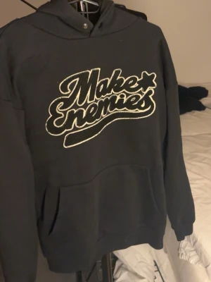 Svart hoodie - Svart hoodie från Make Enemies med stor vit broderad logga på bröstet. Klassisk modell med huva och känguruficka framtill. Perfekt för dig som gillar streetwear och vill sticka ut med en fet print. Mjuk och skön bomullskänsla.