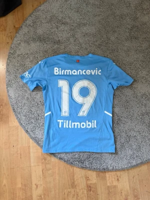 Malmö FF matchtröja Puma ljusblå - Ljusblå Malmö FF matchtröja från Puma med tryck av Birmancevic och nummer 19 på ryggen. Tröjan har klubbmärke, sponsorer och vita detaljer. Perfekt för fotbollsfans som vill visa sitt stöd på läktaren eller planen.