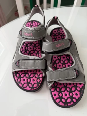 Grå sportiga sandaler med rosa detaljer - Snygga grå sandaler med rosa blommigt mönster på innersulan och justerbara kardborreband. Märket 'Sport' syns på flera ställen. Perfekta för en aktiv stil och riktigt bekväma med mjuk sula och bra stöd.