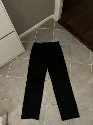 Svarta kostymbyxor Arigato - Säljer ett par klassiska svarta kostymbyxor med raka ben och diskreta bakfickor. Byxorna har en stilren look och passar perfekt till en dressad outfit. Materialet ser ut att vara en blandning av polyester och viskos för en mjuk känsla. Benvidden kan regleras med dragkedja.