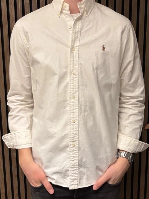 Vit skjorta från Ralph Lauren - Klassisk vit skjorta från Ralph Lauren med button-down krage och broderad logga på bröstet. Skjortan har långa ärmar, knäppning framtill och är tillverkad i ett mjukt bomullstyg. Perfekt för en clean och stilren look.