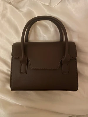 Brun handväska - Säljer super fin helt ny, (med prislapp och allt kvar) brun liten axelremsväska/handväska. Man får med ett band. Den är ungefär i längden som en mobil, något längre💖