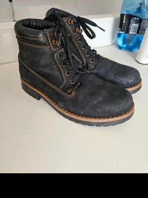 Svarta kängor med kontrastsöm - Säljer ett par svarta kängor i mocka och läder med grov sula och snygga kontrastsömmar i brunt. Klassisk snörning med svarta skosnören och metallöljetter. Perfekta för dig som vill ha en stilren och robust look till hösten eller vintern.