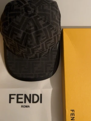Svart keps från Fendi med FF-mönster - Snygg svart keps från Fendi med det klassiska FF-monogrammet över hela kepsen. Kepsen har böjd skärm och är tillverkad i ett tygmaterial som känns lyxigt och trendigt. Perfekt accessoar för att lyfta din streetwear-look.