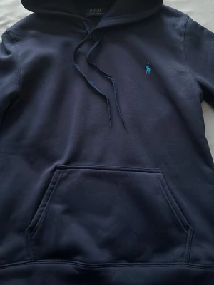 Mörkblå hoodie från Polo Ralph Lauren - Snygg mörkblå hoodie från Polo Ralph Lauren med klassisk huva och dragsnören. Har en stor magficka och den ikoniska blå broderade loggan på bröstet. Tillverkad i mjukt bomullsmaterial som känns riktigt skönt mot huden. Säljer på grund av att den inte passar mig längre fick den som present.