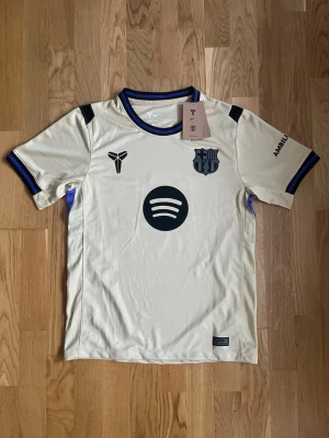 FC Barcelona bortatröja Lamine Yamal - Snygg beige FC Barcelona bortatröja från Nike med Lamine Yamal och nummer 10 på ryggen. Tröjan har svarta detaljer, klubbmärke, Spotify-logga och UNHCR-tryck. Korta ärmar och blåsvarta muddar. Perfekt för dig som älskar fotboll och Barca.