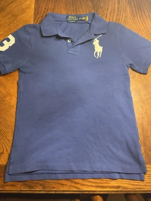 Blå pikétröja från Polo Ralph Lauren - Klassisk blå pikétröja från Polo Ralph Lauren med vit logga på bröstet och siffran 3 på ärmen. Tröjan har krage, knappar vid halsen och korta ärmar. Perfekt för en avslappnad men ändå stilren look.