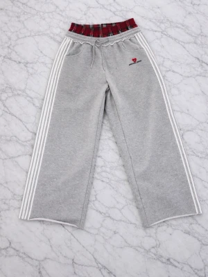 Grå mjukisbyxor Aelfric Eden - Grå mjukisbyxor från Aelfric Eden med vita ränder längs sidorna och en unik röd- och svartmönstrad resår i midjan. Broderad logga med hjärta på vänster sida. Vida ben och dragsko i midjan. Perfekta för en chill och avslappnad stil.