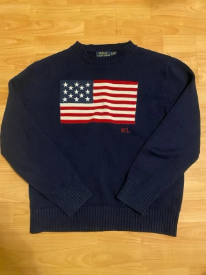 Mörkblå stickad tröja Polo Ralph Lauren M - Mörkblå stickad tröja från Polo Ralph Lauren med amerikansk flagga i rött, vitt och blått på bröstet samt RL-broderi. Tröjan har rund halsringning och ribbade muddar vid ärmslut och nederkant. Perfekt för dig som gillar klassisk preppy stil.
