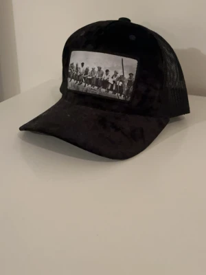 Svart velvet keps med fototryck - Säljer en svart truckerkeps med mesh på sidorna och ett stort fototryck framtill som visar ett klassiskt svartvitt motiv. Kepsen har böjd skärm och justerbar snapback baktill. Perfekt för dig som vill ha en unik och stilren accessoar.Velvet keps😀Aldrig använd.