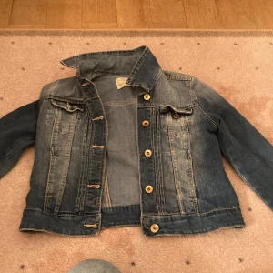 Klassisk blå jeansjacka - Snygg blå jeansjacka med slitningar och kontrastsömmar. Jackan har bröstfickor med lock och knappar framtill i metall. Kortare modell med klassisk krage och raka ärmar. Perfekt att slänga över en hoodie eller t-shirt för en avslappnad look.