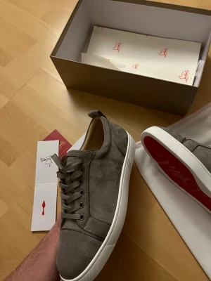 Gråa sneakers  Christian Louboutin - Snygga gråa sneakers i mocka från Christian Louboutin med klassisk röd sula och vita kanter. Skorna har låga snitt, matchande grå skosnören och diskreta detaljer som ger en clean och exklusiv vibe. Perfekta för dig som gillar stilrena och lyxiga sneakers.