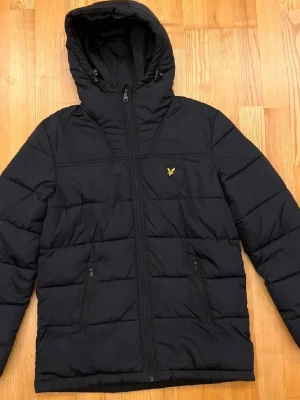 Svart pufferjacka från Lyle & Scott - Svart pufferjacka från Lyle & Scott med huva och dragkedja framtill. Jackan har två sidofickor med dragkedja och det klassiska gula logomärket på bröstet. Quiltad design och varm vaddering, perfekt för kalla dagar.