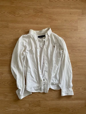 Vit skjorta från Ralph Lauren - Klassisk vit skjorta från Ralph Lauren med broderad logga på bröstet. Skjortan har lång ärm, knappar framtill och en tidlös design. Perfekt för dig som gillar stilrena och enkla plagg. Tillverkad i bomull för en skön känsla. Storlek M/S