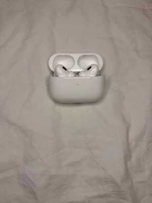 Apple AirPods (gen 2) noise cancellation  - Säljer ett par vita Apple AirPods Pro 2 med trådlöst laddningsetui. Hörlurarna har en stilren och kompakt design i plast och levereras med originalförpackning och tillbehör som instruktioner och extra öronproppar. Perfekta för dig som vill ha grymt ljud och smidig anslutning.