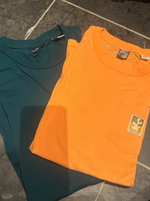 2-pack t-shirts från Scotch & Soda - Två snygga t-shirts från Scotch & Soda, en i mörkgrönt och en i orange med bröstficka och logga. Båda har rund hals och klassisk passform. Perfekta för en avslappnad och stilren look.