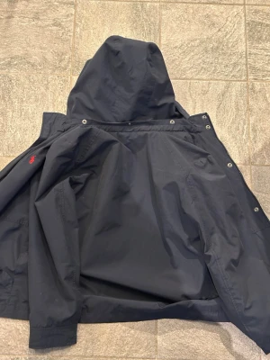 Marinblå vindjacka från Polo Ralph Lauren - Snygg marinblå vindjacka från Polo Ralph Lauren. Luvan går att ta av du stänger jackan via dragkedja eller knappar. Priset går att diskutera