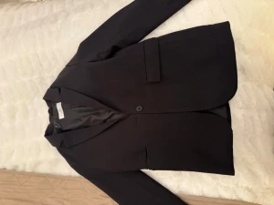 Svart kavaj från H&M - Stilren svart kavaj från H&M med klassisk passform och enkelknäppning. Kavajen har två fickor framtill och är helfodrad med slät insida. Perfekt för en clean och snygg look.